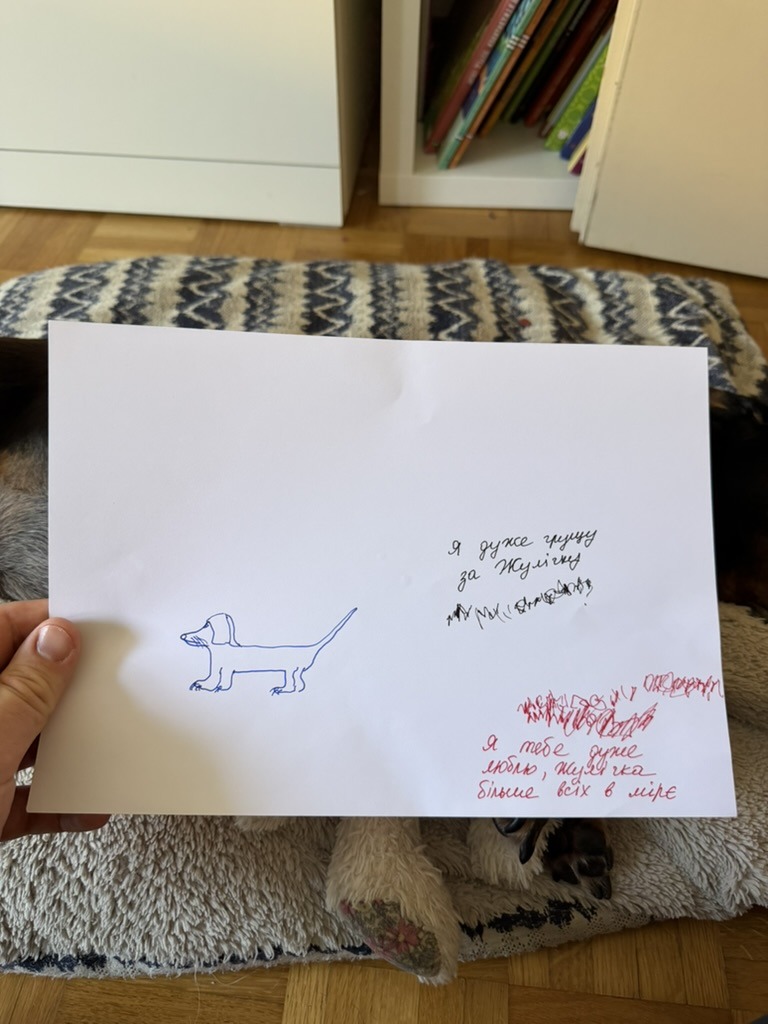 A sheet of white paper held up in one hand. On the left, a line drawing of a dachshund in blue pen. Top right, two lines of adult handwriting in blue with dark scribbles by a child underneath them. Bottom right, red scribbles by a child with three lines of adult handwriting in red underneath.